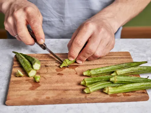 Hände schneiden Okra für ein leckeres Okra-Gericht. Frische grüne Schoten liegen auf einem Holzbrett.