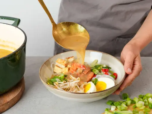 Eine goldene Schöpfkelle gießt eine cremige Brühe über Garnelen, Tofu, Ei und Sprossen in einer Schüssel. Ein köstliches Laksa Rezept wird zubereitet.
