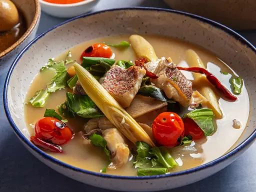 Eine dampfende Schüssel mit **thai Fischsuppe**, garniert mit Kirschtomaten, Kräutern und Zitronengras.