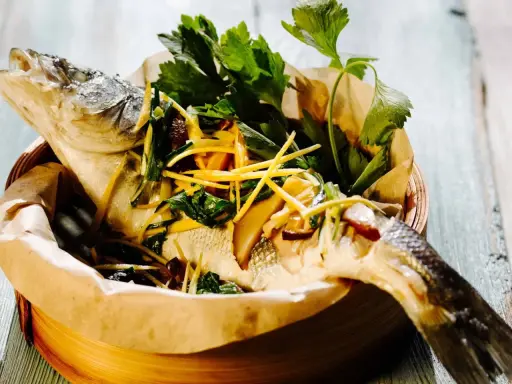Gedämpfter Fisch, asiatisch zubereitet, mit Ingwer, Frühlingszwiebeln und frischen Kräutern in einem Bambuskorb.
