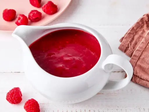 Himbeer coulis in einer weißen Soßenkelle, daneben frische Himbeeren und eine rosa Platte mit weiteren Früchten.