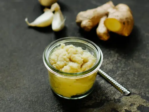 Frisch zubereitete ingwer knoblauch paste in einem Glas, daneben Ingwerwurzel und Knoblauchzehen auf dunklem Untergrund.