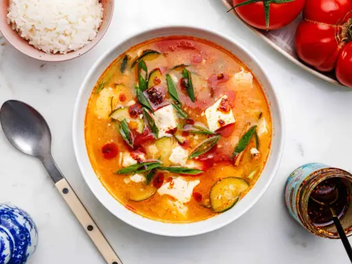 Eine dampfende Schüssel seidentofu suppe mit Zucchini und Frühlingszwiebeln, garniert mit Chiliöl. Daneben eine Schüssel Reis und ein Glas Chiliöl.