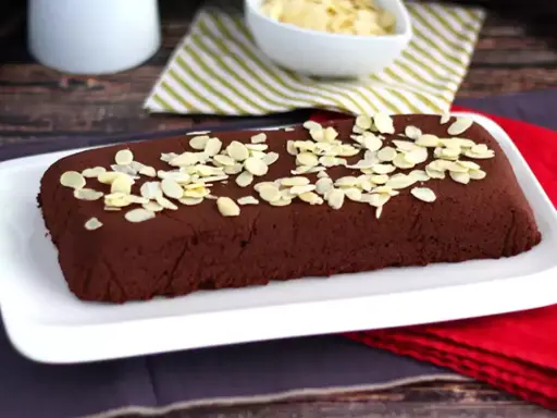 Dunkle, rechteckige marquise au chocolat, bestreut mit Mandelblättchen, auf einem weißen Teller.