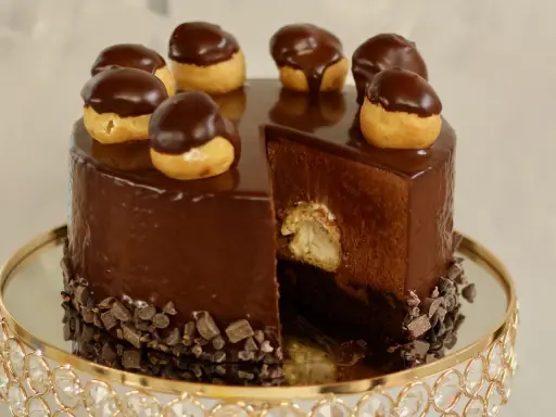 Eine glänzende Schokoladentorte, dekoriert mit kleinen Schokoladen-Profiteroles. Ein Stück ist herausgeschnitten und zeigt die cremige Füllung.