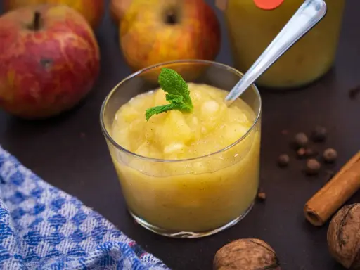 Ein Glas mit hausgemachtem compote de pommes, garniert mit Minze. Äpfel, Zimt und Nüsse im Hintergrund.