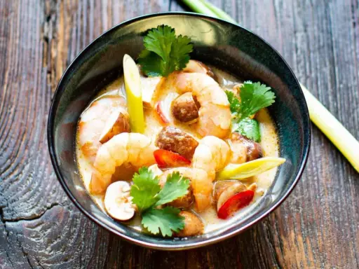 Eine Schüssel mit cremigem Tom Kha Gung, garniert mit Garnelen, Pilzen und Koriander, auf rustikalem Holz.