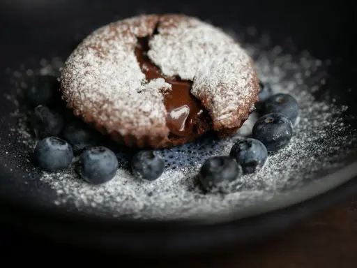 Ein saftiger Moelleux au Chocolat, bestäubt mit Puderzucker, umgeben von frischen Blaubeeren auf einem dunklen Teller.
