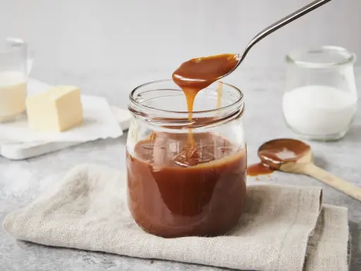 Ein Glas mit glänzender karamellsauce mit milch statt sahne. Ein Löffel hebt die cremige Sauce an.