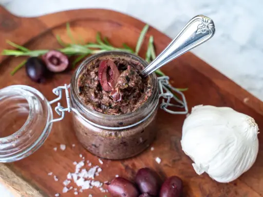 Ein Glas hausgemachte Tapenade, perfekt für Ihr nächstes Tapenade Rezept. Oliven, Knoblauch und Rosmarin auf einem Holzbrett.