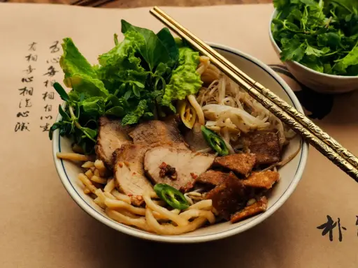 Cao Lau: Nudelschüssel mit Schweinefleisch, Sprossen und Kräutern, garniert mit knusprigen Krusten. Goldene Stäbchen ruhen auf dem Gericht.