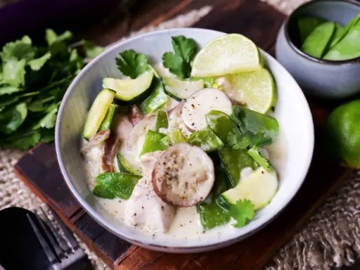 Ein cremiger Thai Green Curry mit Hühnchen, Zucchini, Zuckerschoten und Auberginenscheiben, garniert mit Limettenspalten und Koriander.