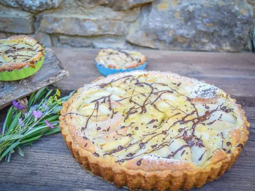 Ein köstlicher Amandine-Kuchen mit Birnenspalten und Schokoladendrizzle, bestäubt mit Puderzucker. Daneben kleine Törtchen und Kräuter.