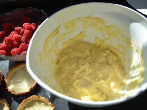 Frische Himbeeren warten neben leeren Tartelettförmchen und einer Schüssel mit cremiger Füllung auf die Fertigstellung der Himbeer Tartelettes.