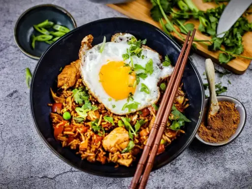 Was ist Nasi Goreng? Ein Teller mit gebratenem Reis, Hühnchen, Erbsen und einem Spiegelei, garniert mit Koriander und Frühlingszwiebeln.