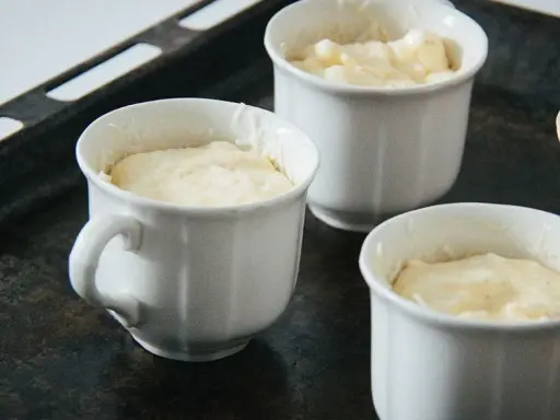 Vier weiße Förmchen mit luftiger Masse für ein Käsesoufflé stehen auf einem Backblech.