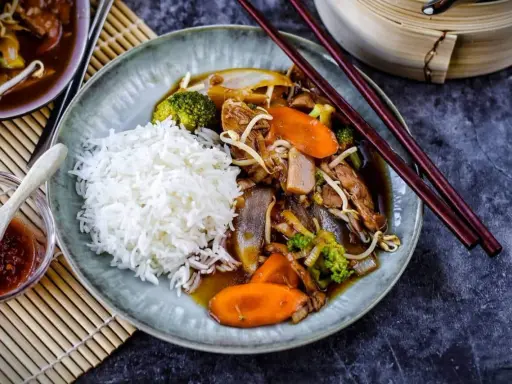Reis mit einem herzhaften Stir-Fry-Gericht, das mit viel Gemüse und Fleisch in einer köstlichen Sauce serviert wird. Dazu gibt es eine kleine Schale mit scharfer Sauce.