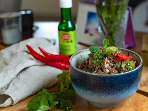 Schüssel mit würzigem Larb Moo, garniert mit frischen Kräutern und Chilis. Daneben liegen rote Chilis und eine Flasche Sauce.