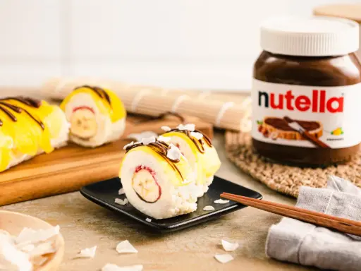 Süße Sushi-Rollen mit Milchreis als Basis, gefüllt mit Banane und Erdbeere, garniert mit gelbem Fruchtpüree, Schokodrizzle und Kokoschips. Daneben eine Nutella-Packung.