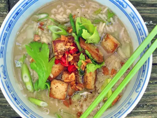 Khao tom, ein herzhafter Reissuppe mit gebratenem Tofu, Frühlingszwiebeln und Kräutern, serviert in einer blauen Schüssel mit Essstäbchen.