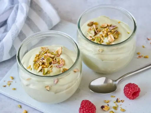 Zwei Gläser mit cremiger Pistazienmousse, garniert mit gehackten Pistazien. Ein Löffel und Himbeeren liegen daneben.