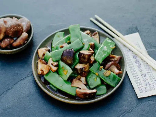 Gebratene Thai Bohnen mit Pilzen in einer dunklen Soße, serviert mit Stäbchen.