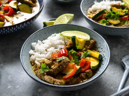 Schüssel mit Reis und rotem Thai Curry mit Rindfleisch, Zucchini und Paprika, garniert mit Limettenspalten und Koriander.