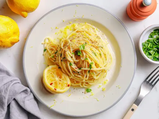 Vegane Pasta mit Zitronenschale und Kräutern, garniert mit einer Zitronenhälfte und frisch gemahlenem Pfeffer.