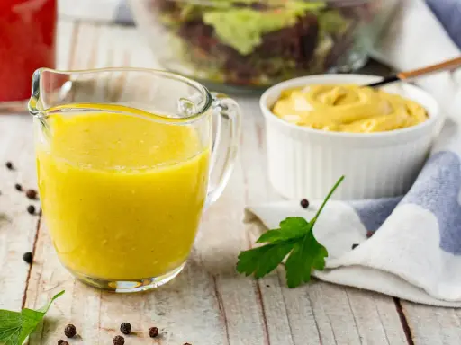 Gelbe Vinaigrette in einem Glasbehälter, bereit für einen frischen Salat. Daneben eine Schüssel Senf und ein Salat im Hintergrund.