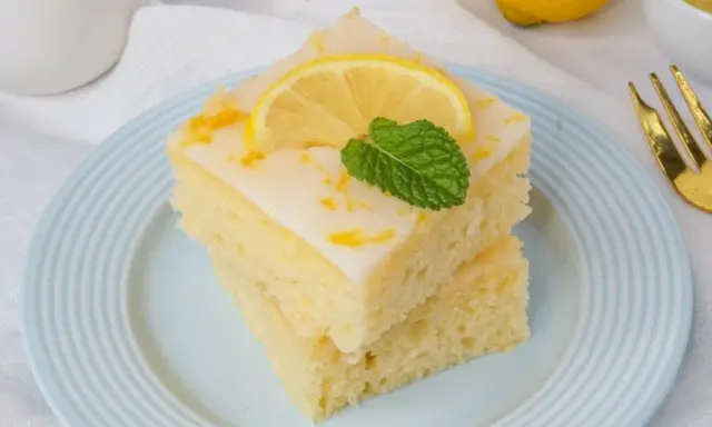 Zwei saftige Stücke weltbester Zitronenkuchen mit Joghurt, garniert mit Zitrone und Minze, auf einem blauen Teller.