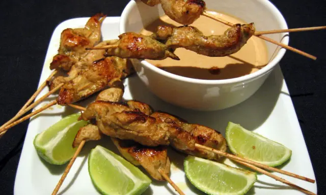 Saftige sate spiesse mit Erdnusssauce und Limettenspalten auf einem weißen Teller.