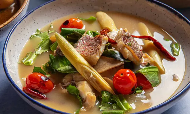 Eine dampfende Schüssel mit **thai Fischsuppe**, garniert mit Kirschtomaten, Kräutern und Zitronengras.
