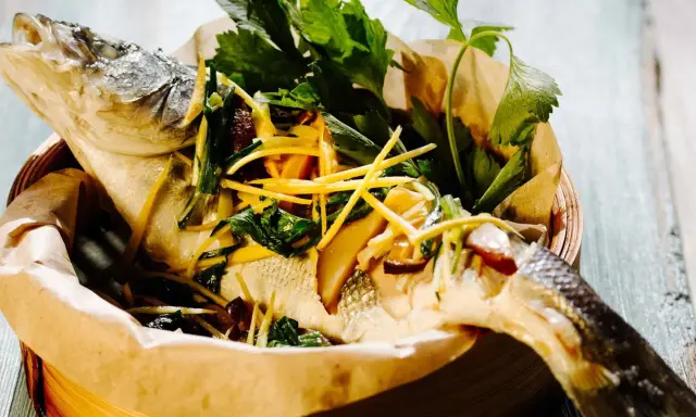 Gedämpfter Fisch, asiatisch zubereitet, mit Ingwer, Frühlingszwiebeln und frischen Kräutern in einem Bambuskorb.