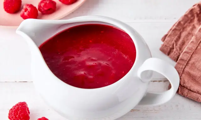 Himbeer coulis in einer weißen Soßenkelle, daneben frische Himbeeren und eine rosa Platte mit weiteren Früchten.