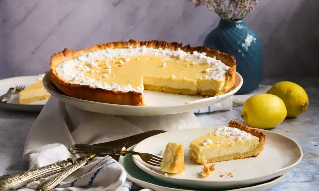 Eine köstliche Zitronen-Tarte, bestäubt mit Puderzucker, steht auf einem Kuchenständer. Ein Stück liegt auf einem Teller, daneben zwei frische Zitronen.