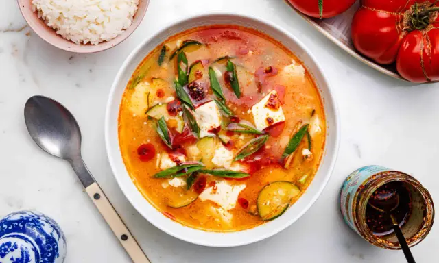 Eine dampfende Schüssel seidentofu suppe mit Zucchini und Frühlingszwiebeln, garniert mit Chiliöl. Daneben eine Schüssel Reis und ein Glas Chiliöl.