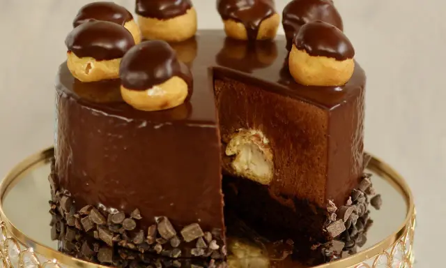 Eine glänzende Schokoladentorte, dekoriert mit kleinen Schokoladen-Profiteroles. Ein Stück ist herausgeschnitten und zeigt die cremige Füllung.