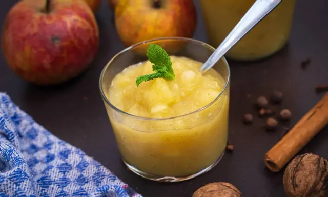 Ein Glas mit hausgemachtem compote de pommes, garniert mit Minze. Äpfel, Zimt und Nüsse im Hintergrund.