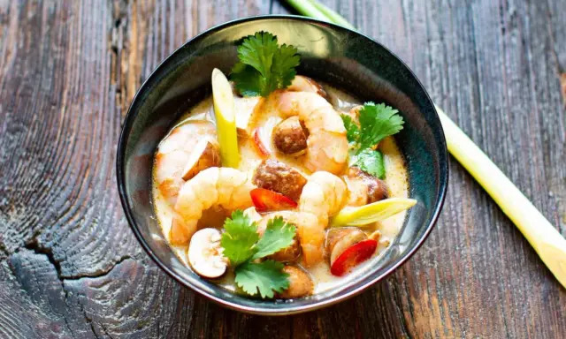 Eine Schüssel mit cremigem Tom Kha Gung, garniert mit Garnelen, Pilzen und Koriander, auf rustikalem Holz.