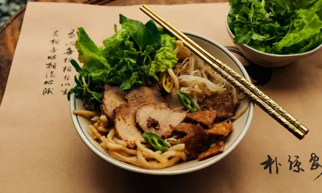 Cao Lau: Nudelschüssel mit Schweinefleisch, Sprossen und Kräutern, garniert mit knusprigen Krusten. Goldene Stäbchen ruhen auf dem Gericht.