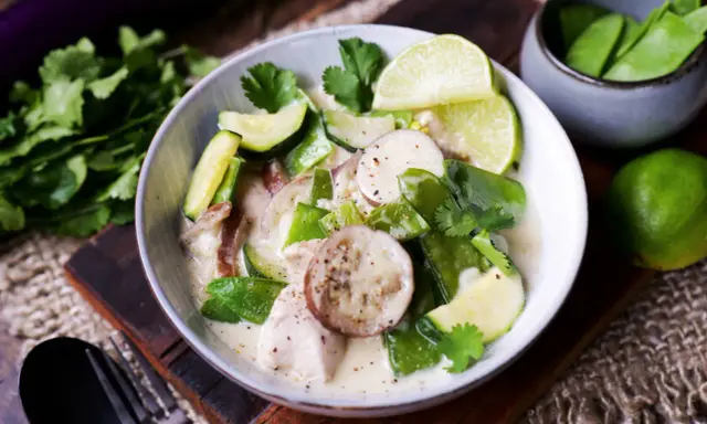 Ein cremiger Thai Green Curry mit Hühnchen, Zucchini, Zuckerschoten und Auberginenscheiben, garniert mit Limettenspalten und Koriander.