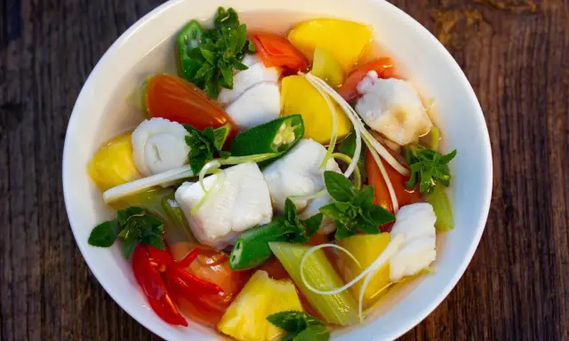 Ein Schüssel Canh Chua mit Fischstücken, Ananas, Tomaten, Okra und Kräutern.