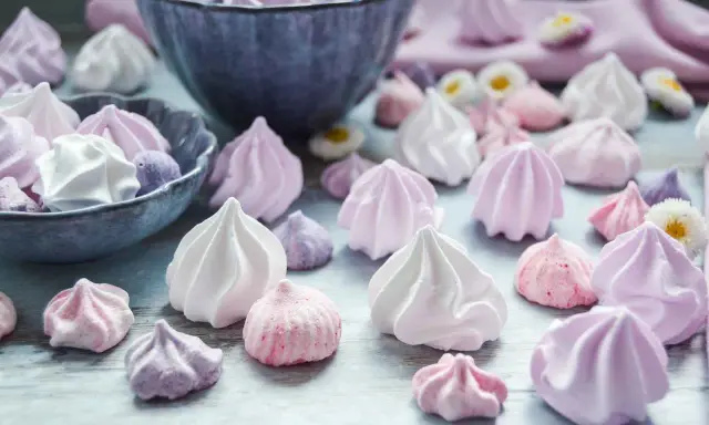 Zuckergebäck, was ist Meringue? Zarte, luftige Baisers in Rosa-, Lila- und Weißtönen, einige in einer Schüssel, andere verstreut.