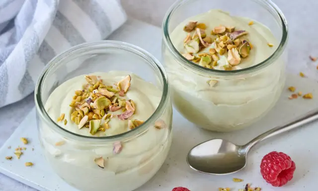Zwei Gläser mit cremiger Pistazienmousse, garniert mit gehackten Pistazien. Ein Löffel und Himbeeren liegen daneben.