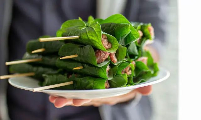 Bo la lot, ein vietnamesisches Gericht, auf einem Teller präsentiert. Fleisch ist in grüne Blätter gewickelt und mit Holzspießen fixiert.