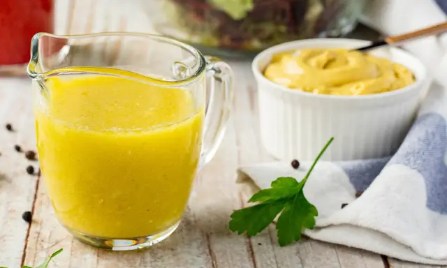 Gelbe Vinaigrette in einem Glasbehälter, bereit für einen frischen Salat. Daneben eine Schüssel Senf und ein Salat im Hintergrund.