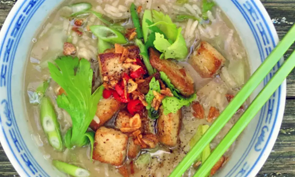 Khao tom, ein herzhafter Reissuppe mit gebratenem Tofu, Frühlingszwiebeln und Kräutern, serviert in einer blauen Schüssel mit Essstäbchen.