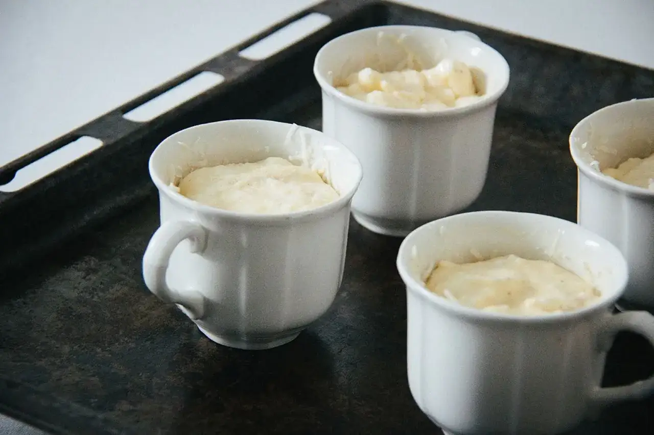 Vier wei&szlig;e F&ouml;rmchen mit luftiger Masse f&uuml;r ein K&auml;sesouffl&eacute; stehen auf einem Backblech bereit.