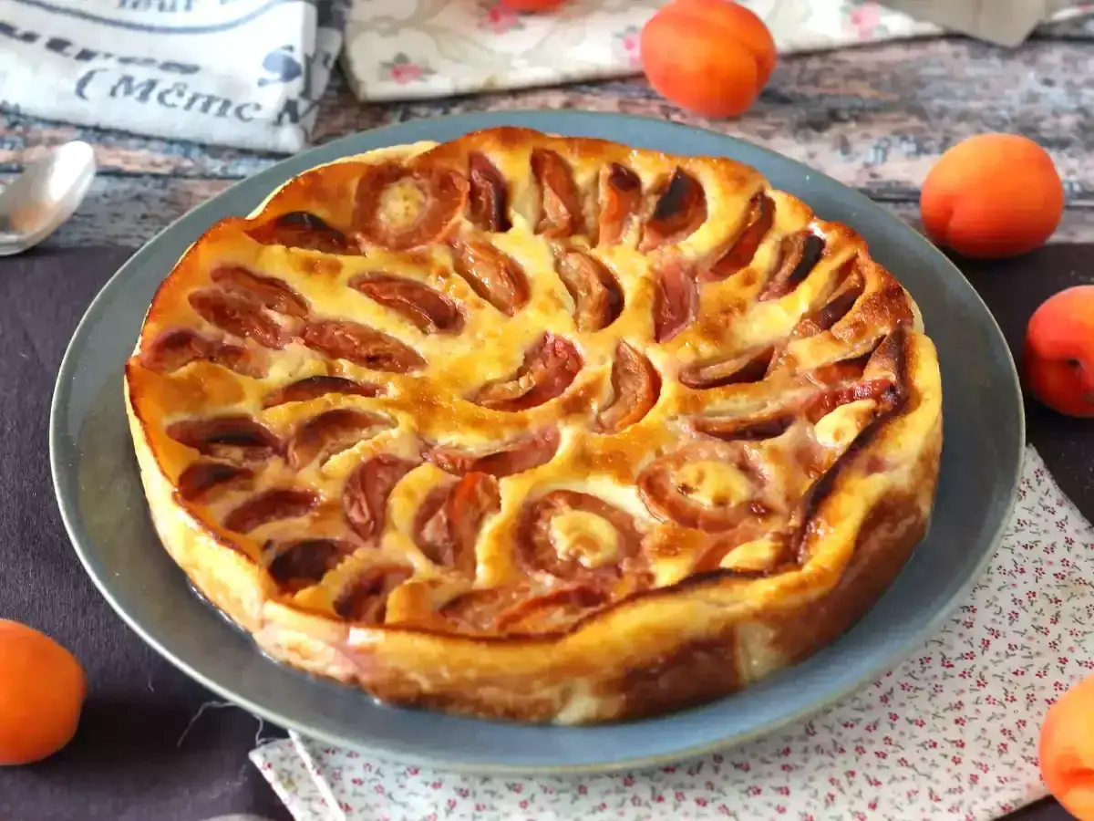 Ein goldbrauner Aprikosen Clafoutis auf einem blauen Teller, umgeben von frischen Aprikosen.