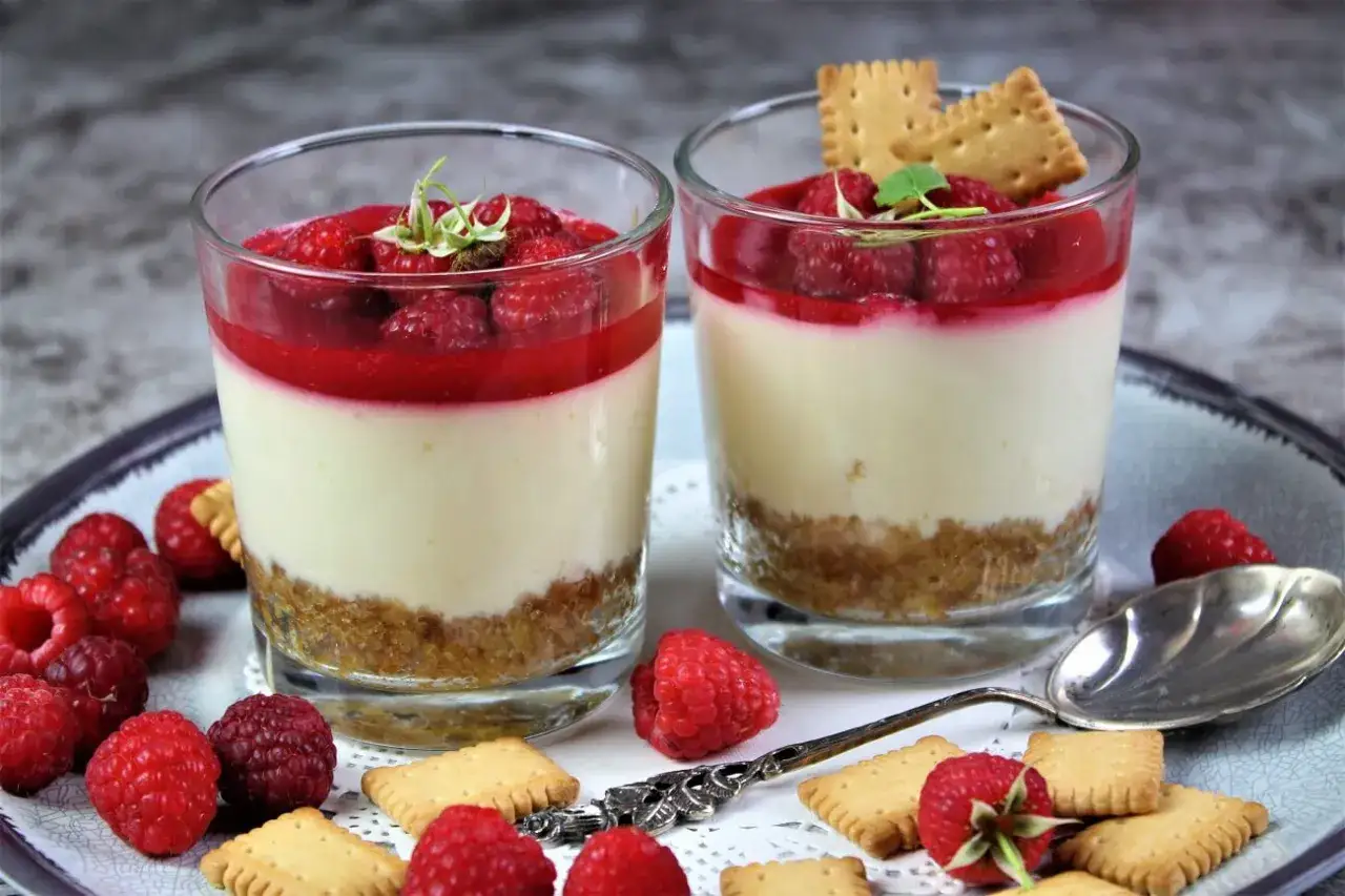 Zwei Gläser mit cremigem Fromage Blanc Dessert, garniert mit Himbeeren und Keksen.